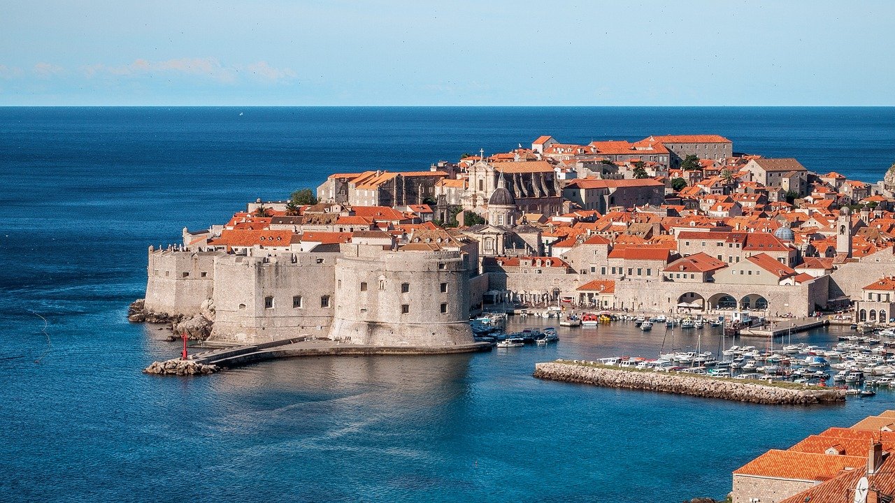 Vue sur les remparts médiévaux de Dubrovnik, majestueux et bien préservés, encerclant la ville historique avec le port animé en premier plan, et les toits de tuiles rouges emblématiques s’étendant à l’horizon