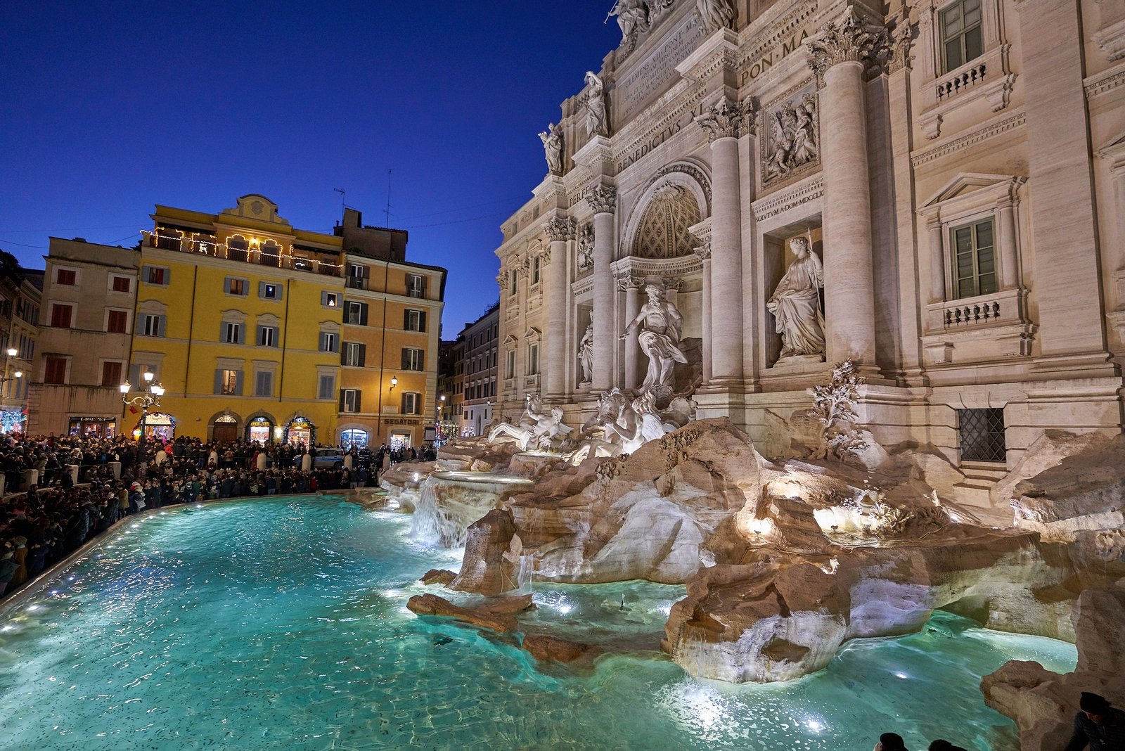 Vue sur la fontaine de Trévi illuminée le soir, mettant en valeur ses sculptures baroques majestueuses, entourée par l'ambiance romantique des lumières de Rome