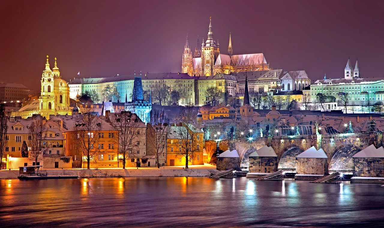 Vue nocturne sur les bâtiments de Prague sous la neige, avec le château de Prague illuminé en arrière-plan, créant une ambiance hivernale féerique