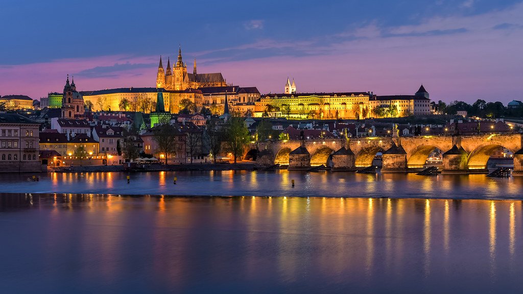 Vue panoramique sur la ville de Prague, illuminée au coucher du soleil, avec le pont Charles en premier plan et le château de Prague en arrière-plan, offrant un paysage emblématique de la capitale tchèque