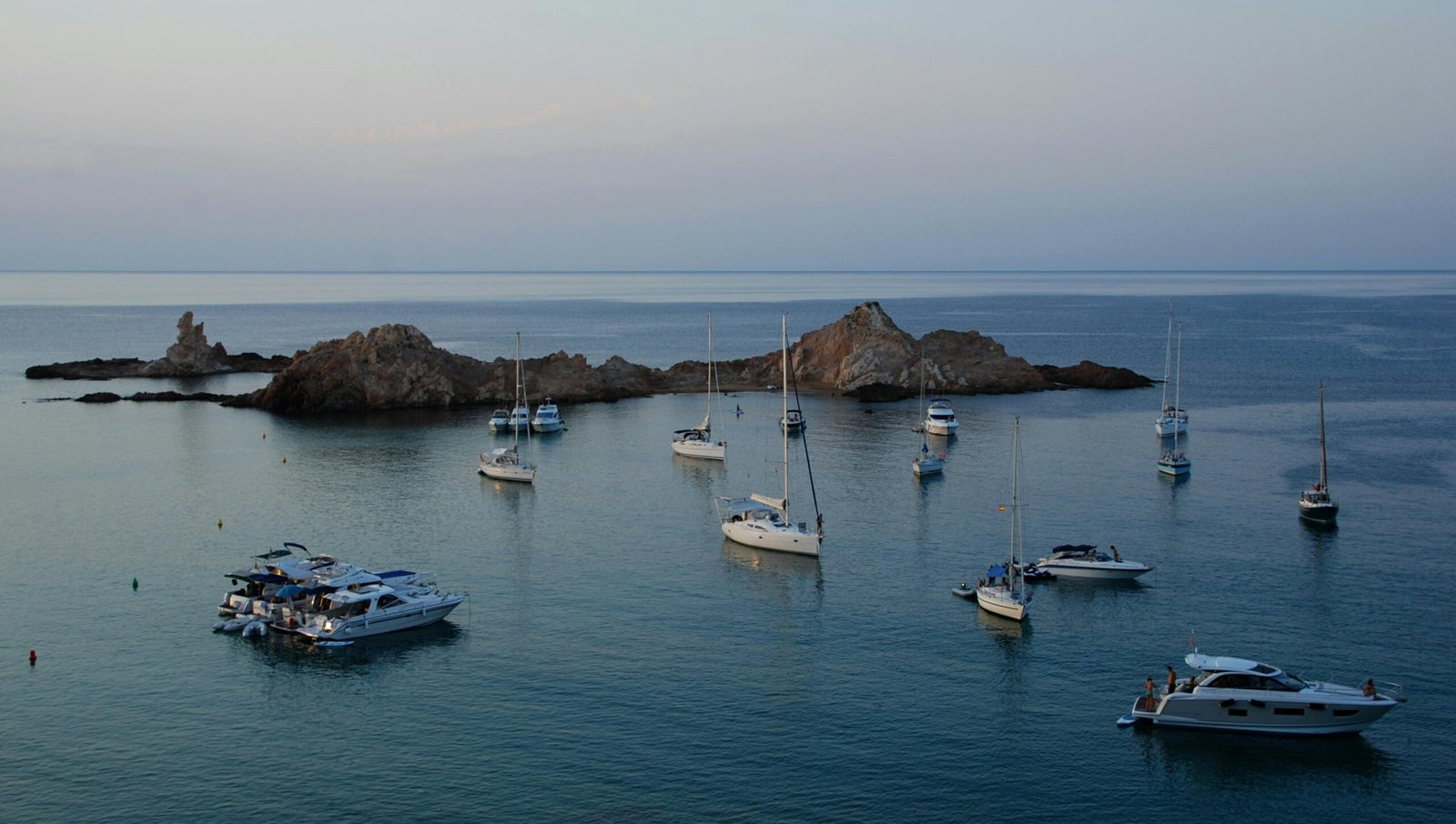 Coucher de soleil sur la côte de Minorque avec plusieurs bateaux amarrés, baignant dans une mer paisible aux reflets dorés et orangés